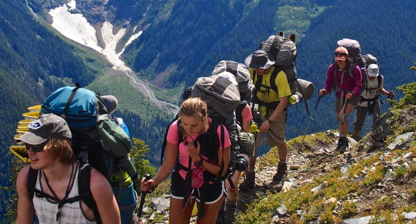 A group of students wearing backpacks hike along a trail high above a mountainous valley containing snow.